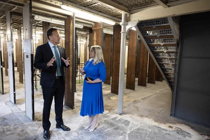 Taoiseach Leo Varadkar, wearing a dark suit and blue tie, is standing and speaking closely with NLI Director Audrey Whitty, wearing a long blue dress, in the West Wing of the NLI. The concrete floors are bare, with evidence of building works. The pair stand among empty book stacks, no longer holding books or shelving. To the right, an old staircase is visible.