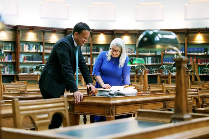 A bright photograph featuring Taoiseach Leo Varadkar, smiling, wearing a dark suit and light coloured tie, with Director of NLI, Audrey Whitty. The pair are smiling as they lean over a booklet which is perched on a wooden table in the middle of the NLI's reading room. Bookshelves adorn the walls in the background of the image, and each desk has a wooden lamp with a green reading lampshade. 