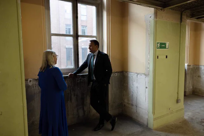 Dr. Audrey Whitty, Director of the NLI (wearing a long blue dress) has her back to the camera and is visible in conversation with Taoiseach Leo Varadkar, who is leaning against an old ledge inside the NLI's west wing building, which is vacant of collections. Bare concrete floors can be seen, and crumbling concrete on the walls. Evidence of some building works can be observed. The window peers out onto Kildare street.