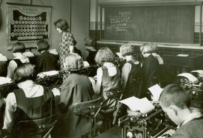 Monochrome photo of predominantly female students learning to type on typewriters while a teacher stands by