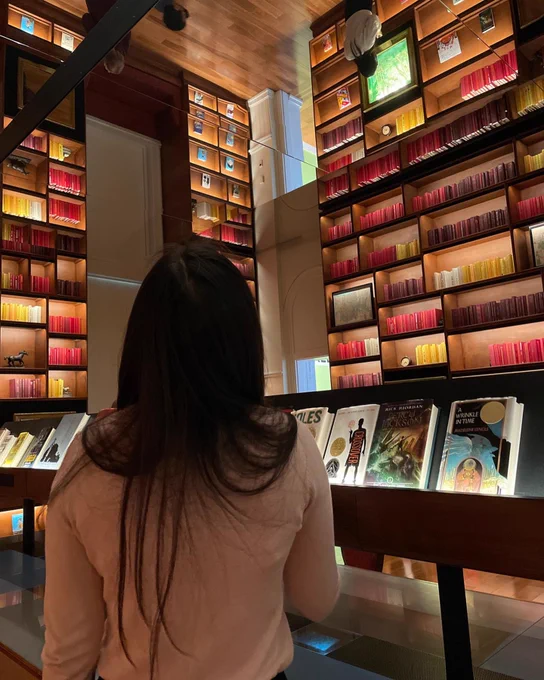 A woman standing in Planet Word’s Library with her back to the camera. In front of her are tall bookshelves reaching up to the ceiling, which is mirrored to make the bookshelves look twice as tall,  and tables with books on display.