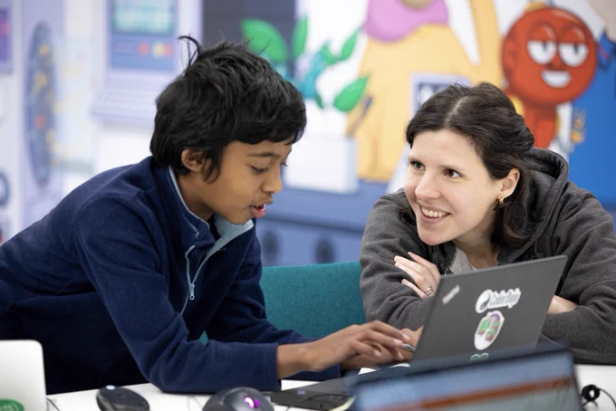 Child coding on a laptop seated beside a mentor