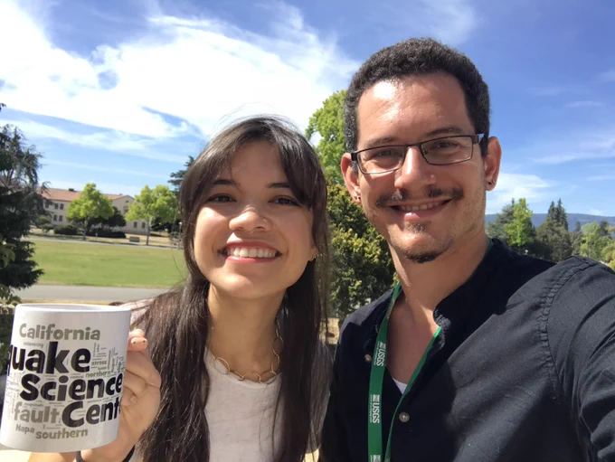 Image shows a smiling young woman and smiling young man taking a selfie with green trees and grass behind them, as well as some low-lying structures and blue skies. The woman is holding up a coffee mug that says USGS California Earthquake Science Center.