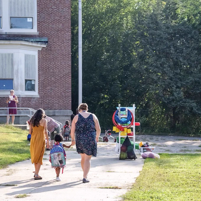 Two Ashland Preschool teachers walk a student wearing an oversized backpack along the sidewalk to the school