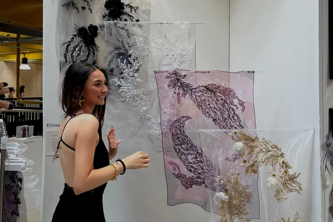 A woman with black hair and a black dress smiles by embroidered art work.