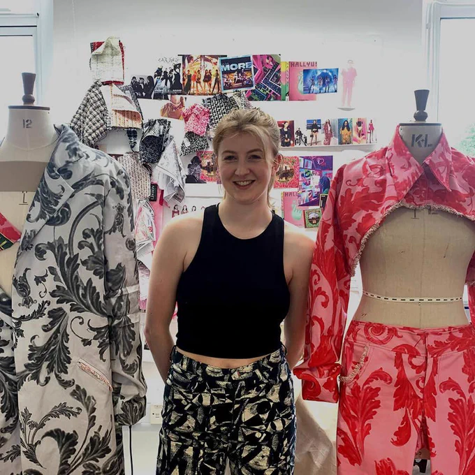 A woman with blonde hair smiles between two colourful outfits on display.