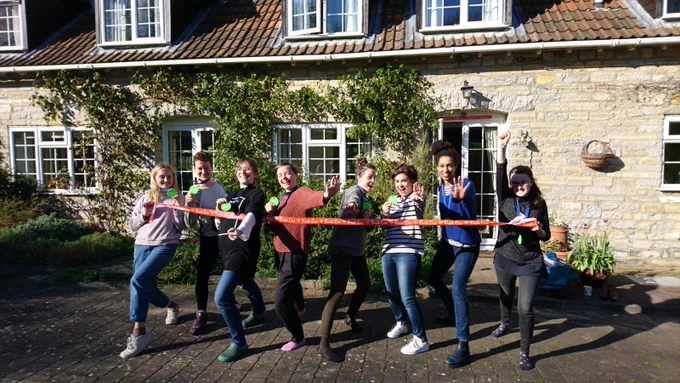 8 people stand in a line in front of a house, some holding their hands out in front of them, all holding red 'finish line' tape.