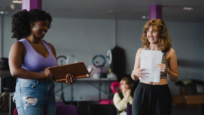 Jada-Li Warrican and Rosa Hesmondhalgh in rehearsals for We Could All Be Perfect. Photo by Becky Payne. Two women laugh and smile. They stand next to one another in a rehearsal room. One holds an open book and the other clutches a sheet of paper to their chest.