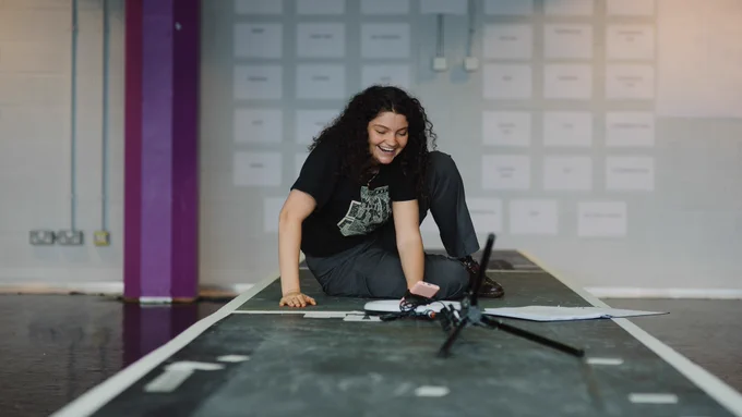 Alice Walker in rehearsals for We Could All Be Perfect. Photo by Becky Payne. A young woman wearing back sits on a raised platform in front of a phone on a holder. They laugh as they read from a script on the floor in front of them.