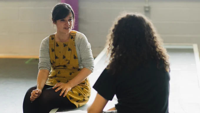 Director Ruby Clarke in rehearsals for We Could All Be Perfect. Photo by Becky Payne. Two women sit on the edge of a raised platform and are in conversation. One wears black and the other wears a yellow pinafore.