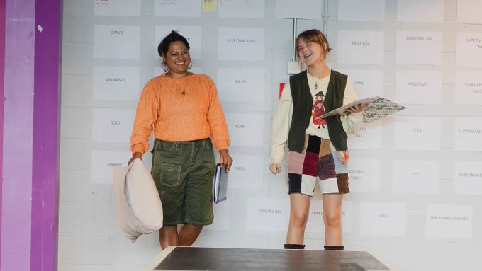 Anshula Bain and Heather Forster in rehearsals for We Could All Be Perfect. Photo by Becky Payne. Two young women laugh and smile, standing at the end of a long, thin platform. One holds a cushion and a folder in their hands, the other holds an open folder as if reading from it. Behind are lots of pieces of paper stuck across the wall with singular words on them. 