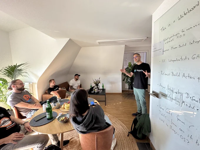 Vinai standing in a living room kitchen in front of  people sitting on sofa and a table. Subtle sunlight in the room. A glass door on the right part of the picture is used as a whiteboard with some topic proposals for the hackathon.