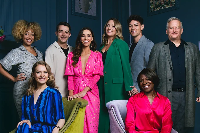 A photo showing a group of eight people grouped together against the wall of a room, dressed in bright, warm clothing. 
