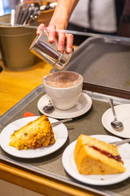 Two slices of cake on two small plates on a tray with a hot drink in a cup on a saucer. A hand is garnishing the drink with chocolate sprinkles. 