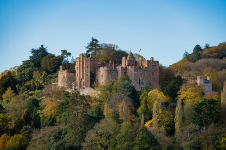A photograph of Dunster Castle taken from a distance. The castle is perched on a wooded hill, where the leaves on the trees are beginning to turn to autumnal colours. 