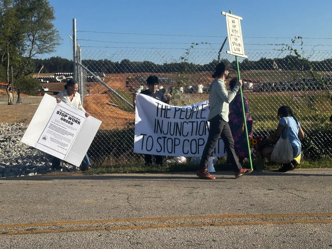 A banner reads “the people’s injunction to stop cop city” held up in front of a construction site. Five people are visible setting up the signage for their protest.