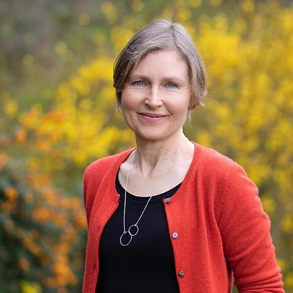 Alice is standing against a blurred green background. She has kind blue eyes and grey/brown hair. she is wearing a black top with a longsleeved red cardigan. 
