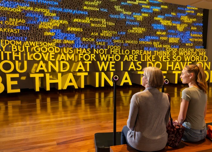 Planet Word visitors sitting in front of microphones with glowing rainbow rings with three-dimensional words lit up in blue and yellow on Planet Word’s Word Wall in the background.