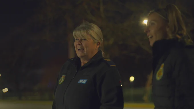 Mandy standing outside at night and wears her paramedic uniform. There is grass and trees behind her. She is standing with her partner.
