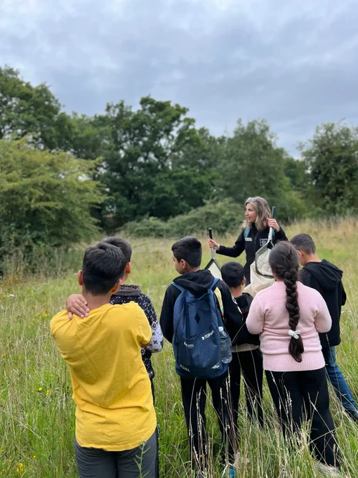 A member of the NWT education team is showing children around the nature reserve. There is tall green and yellow grasses and trees in the distance. 