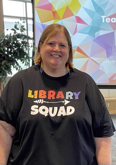 Nicole@is wearing a black teacher with rainbow writing that say Library Squad.