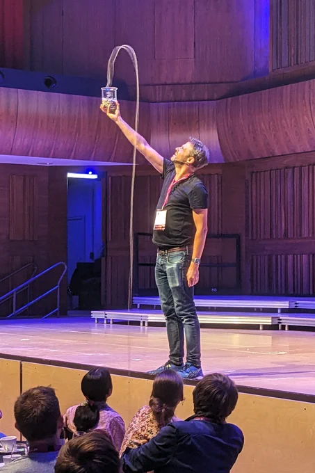 Steve Mould demonstrating 'The Mould Effect'. He's standing on stage, holding a glass beaker containing a string of metallic beads. As the string pours out of the glass, it climbs into the air forming an arc above the beaker.