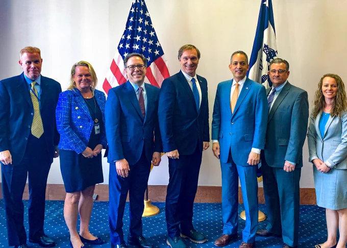 Leaders from the Department of Commerce (Deputy Secretary Don Graves), National Telecommunications and Information Administration (Administrator Alan Davidson, April Delaney, and Mike Dame), and FirstNet Authority (Executive Director and CEO Joe Wassel, Board Chair Richard Carrizzo, April Ward) stand in front of flags 