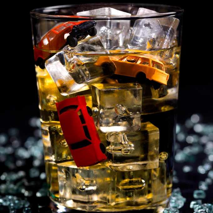 Photograph of a glass with ice, amber-colored liquid, and toy cars.