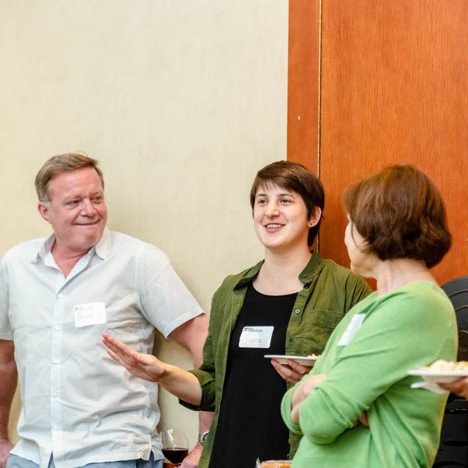 A faculty member and two students stand indoors together conversing.