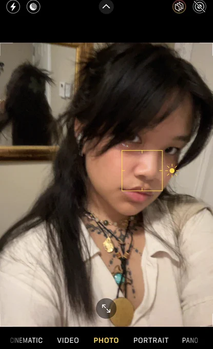 a screenshot from the camera app of anata laylay posing for the camera. the focus sunshine button is focused on their nose. the photo is slightly blurry from motion. they’re wearing a beige button up with four necklaces on top of their chest tattoo.