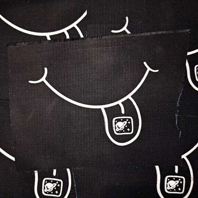 A black fabric patch with white ink that depicts a smiling mouth with it's tongue out
 on the tongue is a square that has a galaxy inside it.