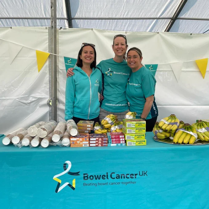 Image of three members of Bowel Cancer UK staff stood smiling for a photo. They are in a marquee and stood behind a stall table full of food for runners.