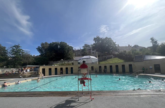A lifeguard watches over a bright blue crescent shaped pool under a sunny sky
