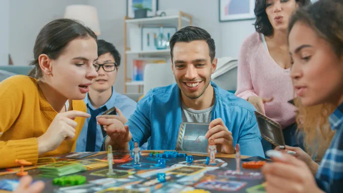 Five people huddled around a table playing a board game. It looks to involve a space ship, tokens, dice, cubes, and cards. They appear to be sitting in a living room. The hand of a sixth person is in the bottom-left.
