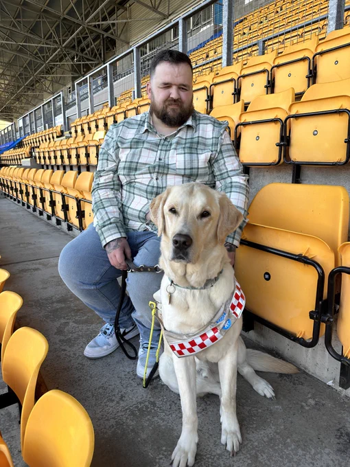 Jon sitting in the stands at McDiarmid Park, Sam the guide dog is sitting next to him wearing his guide dog harness and looking at the camera