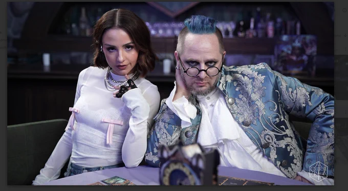 Aimee Carrero and Talieisn Jaffe sit at a table with the Queen by Midnight Spread, Aimee iwearing a white long sleeved top and pearl necklaces and Talieisn is wearing a high caollared white frill shirt and a blue and white brocade and paisely verailles style overcoat, and round glasses 
