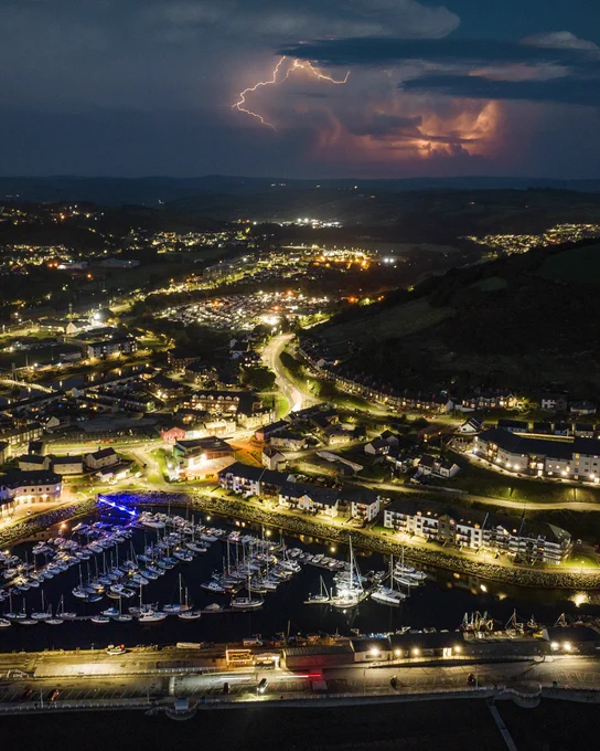 An aerial shot of the southern end of Aberystwyth at night, showing the marina. Multiple lighting strikes can be seen over the hills in the distance.
