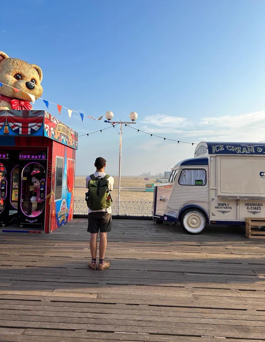 a person in a hat is looking onto the sand from a wooden pier, flanked by two structures. On their left a red amusement block with a large teddy bear on top and to their right, a vintage Citroen food van