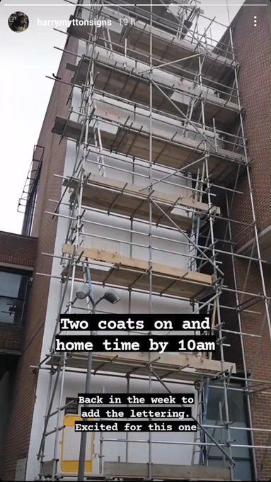 A screenshot of a short film that views a high wall from bottom to top. This shot shows most of the tall, thin wall. It has scaffold in front of it and had been painted white, unlike the dark reddy bricks of the other facades. The screenshot has text over it, "Two coats on and home by 10am" and "Back in the week to add the lettering".

Image: Harry Mytton.