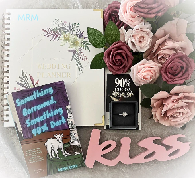 A paperback copy of the book with a wedding planner, roses, chocolate bar, engagement ring, and the word kiss