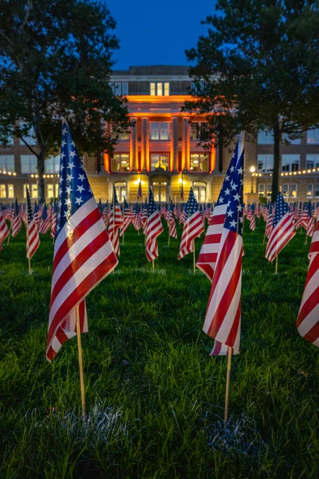 Ground-level photo of the Bowen-Thompson Quadrangle at night. In view, there are hundreds of the nearly 3,000 American flags in the lawn. University Hall is lit orange in the background, the sky is a dark bright blue at dusk. 