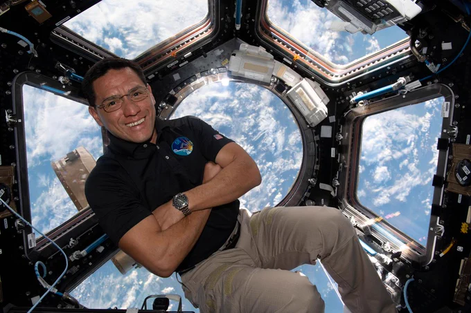 Frank Rubio floats in the space station's cupola. His arms are crossed and he smiles at the camera. Earth is in the background, showing its scattered clouds. Credit: NASA