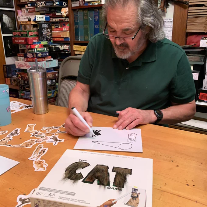 Tim drawing a movie image in The Cat Game on a wooden table next to the game box, cat images on a wooden table with games stacked behind him.