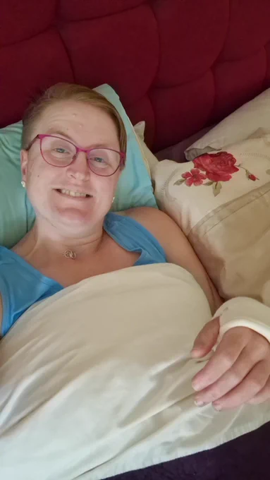 Photo of Ruth lying in bed, a blue pillow behind her and a red headboard. She's wearing a blue vest top and smiling