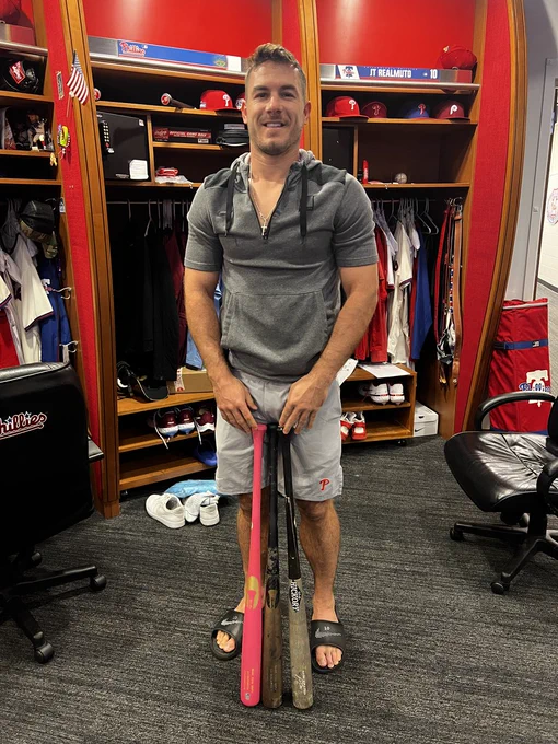 Photo of J.T. Realmuto smiling in the Phillies clubhouse with 3 of his bats.  He is wearing a gray, short sleeve, quarter zip hoodie, gray shorts and black Nike slides.
