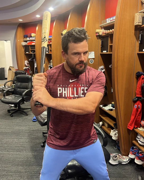 Photo of Kyle Schwarber making a funny face while holding one of his bats.  He is wearing the Phillies powder blue pants with a dark red Phillies t-shirt.  He's standing in the Phillies clubhouse.