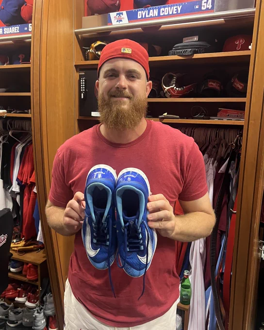 Photo of Dylan Covey in the Phillies clubhouse.  He is holding a pair of blue Nike cleats while smiling.  He's wearing white pants, a red t-shirt and a red hat backwards.