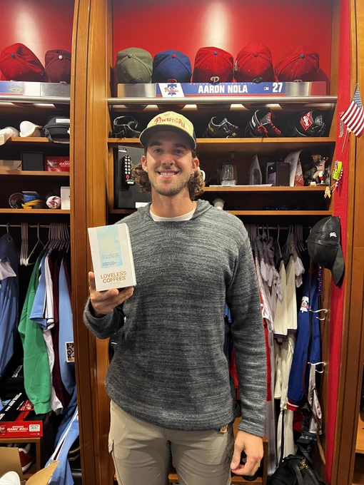 Photo of Aaron Nola in the Phillies clubhouse.  He is wearing a gray sweater, khaki pants and a hat.  He is holding a coffee blend by the company "Loveless Coffees".