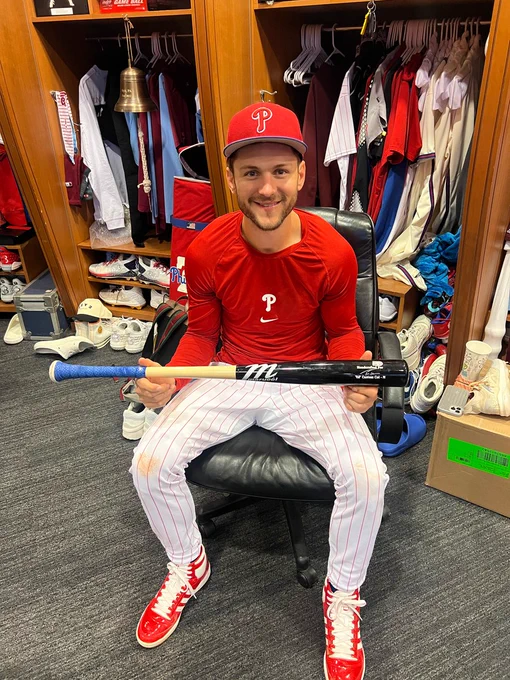 Photo of Trea Turner in the Phillies clubhouse holding one of his bats.  He is wearing a red long sleeve Phillies shirt, red and white pinstripe baseball pants, a red Phillies hat and red sneakers.