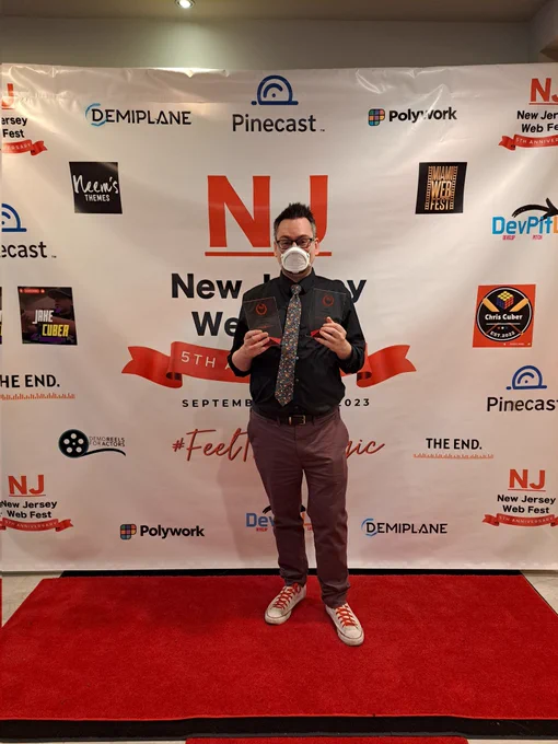 Red Carpet photo of Jeff at NJ Web Fest, holding the awards for Best Series Premise in an Actual Play and Best Anthology. He's wearing a black dress shirt, floral tie, burgundy pants, white Chucks with red laces, and a FloMask.